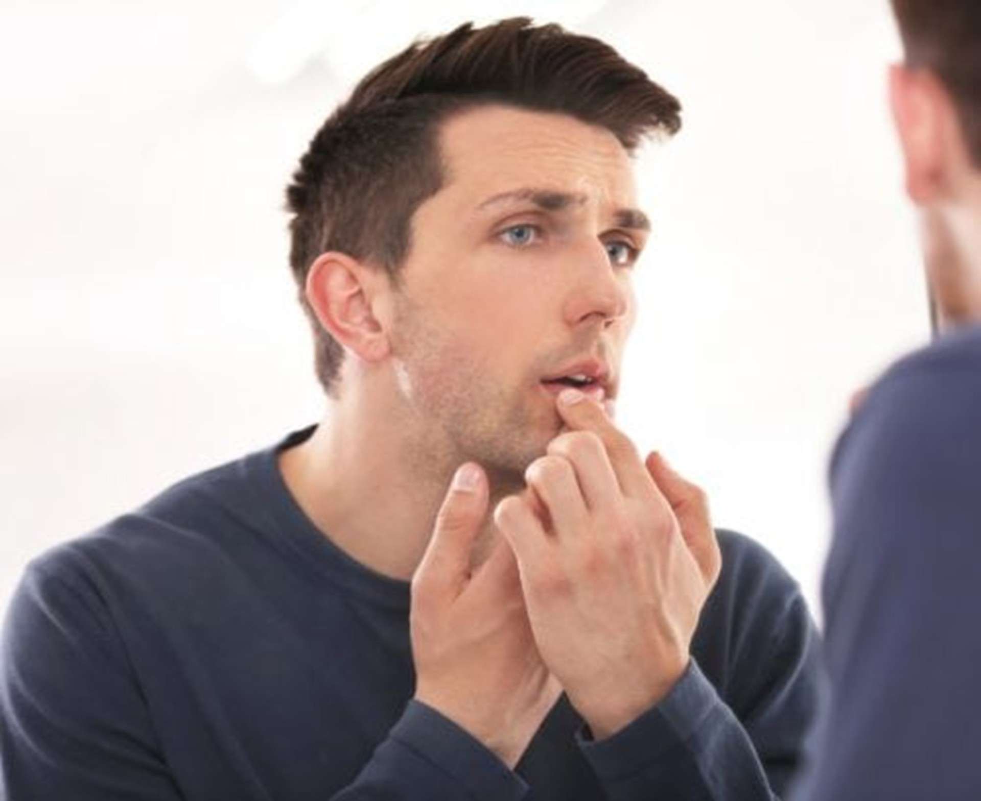A man standing in front of a mirror, touching his lower lip with a concerned expression, possibly examining a cold sore on his mouth
